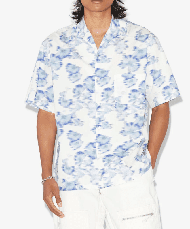 Une personne porte une chemise à manches courtes avec un motif floral bleu et blanc et un pantalon blanc avec des détails de fermeture éclair. La personne a les cheveux longs et se tient debout sur un fond blanc uni.