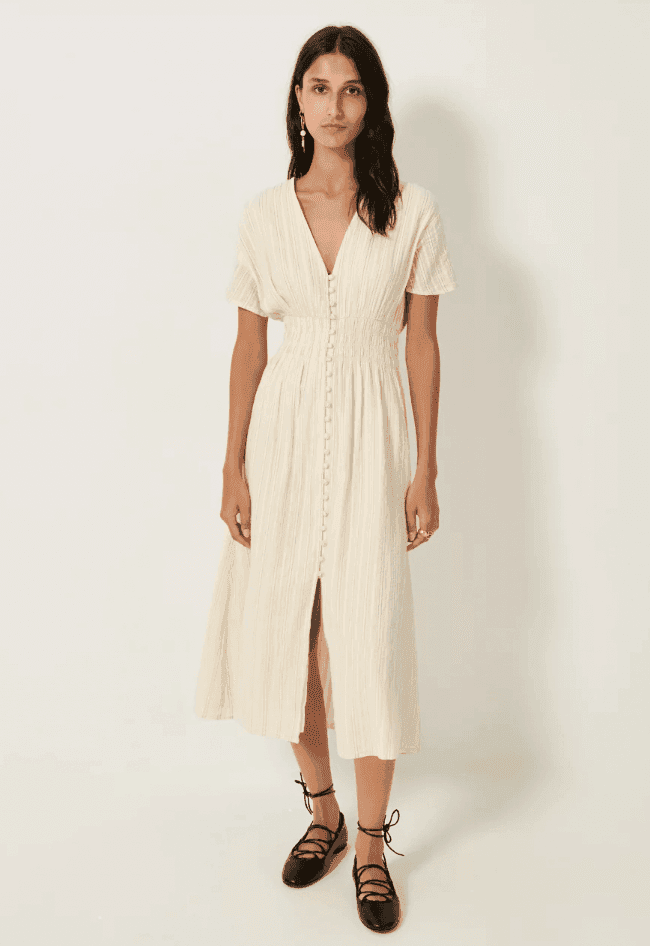 Femme portant une robe midi boutonnée de couleur crème à manches courtes et à taille froncée. Elle a de longs cheveux bruns, se tient debout sur un fond blanc uni et porte des chaussures plates noires à lacets à la cheville.