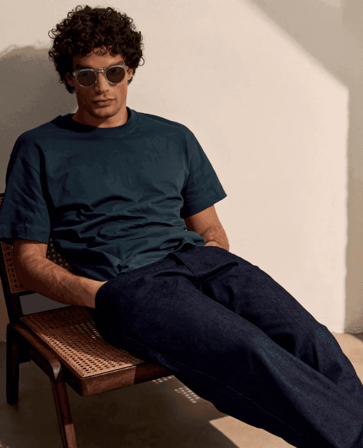 Une personne aux cheveux bouclés, portant des lunettes de soleil, un t-shirt vert foncé et un pantalon foncé, est assise sur une chaise tressée contre un mur de couleur claire. Elle a une posture détendue, les mains dans les poches.