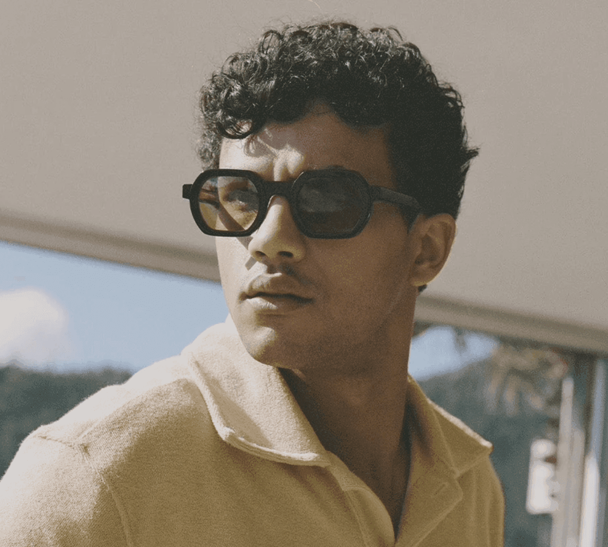 Une personne aux cheveux bouclés, portant des lunettes de soleil foncées et une chemise à col jaune clair, regarde sur le côté. L'arrière-plan montre un ciel clair et un feuillage flou.