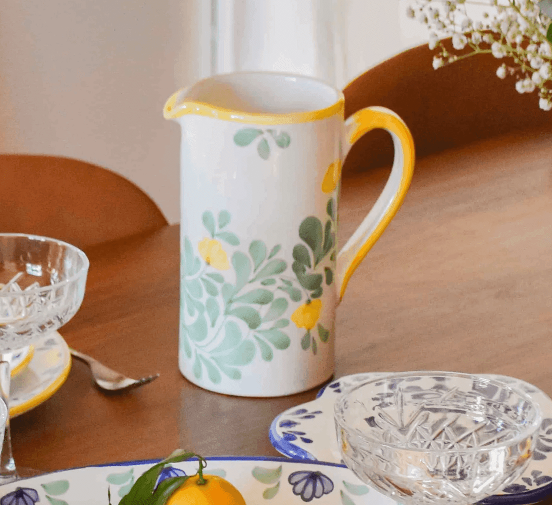 Une cruche en céramique avec une anse et un bec verseur jaunes, ornée de feuilles vertes et de fleurs jaunes, est posée sur une table en bois. À côté se trouvent des bols en verre, une assiette à motif floral, une cuillère et un petit fruit orange. Des fleurs blanches sont à l'arrière-plan.