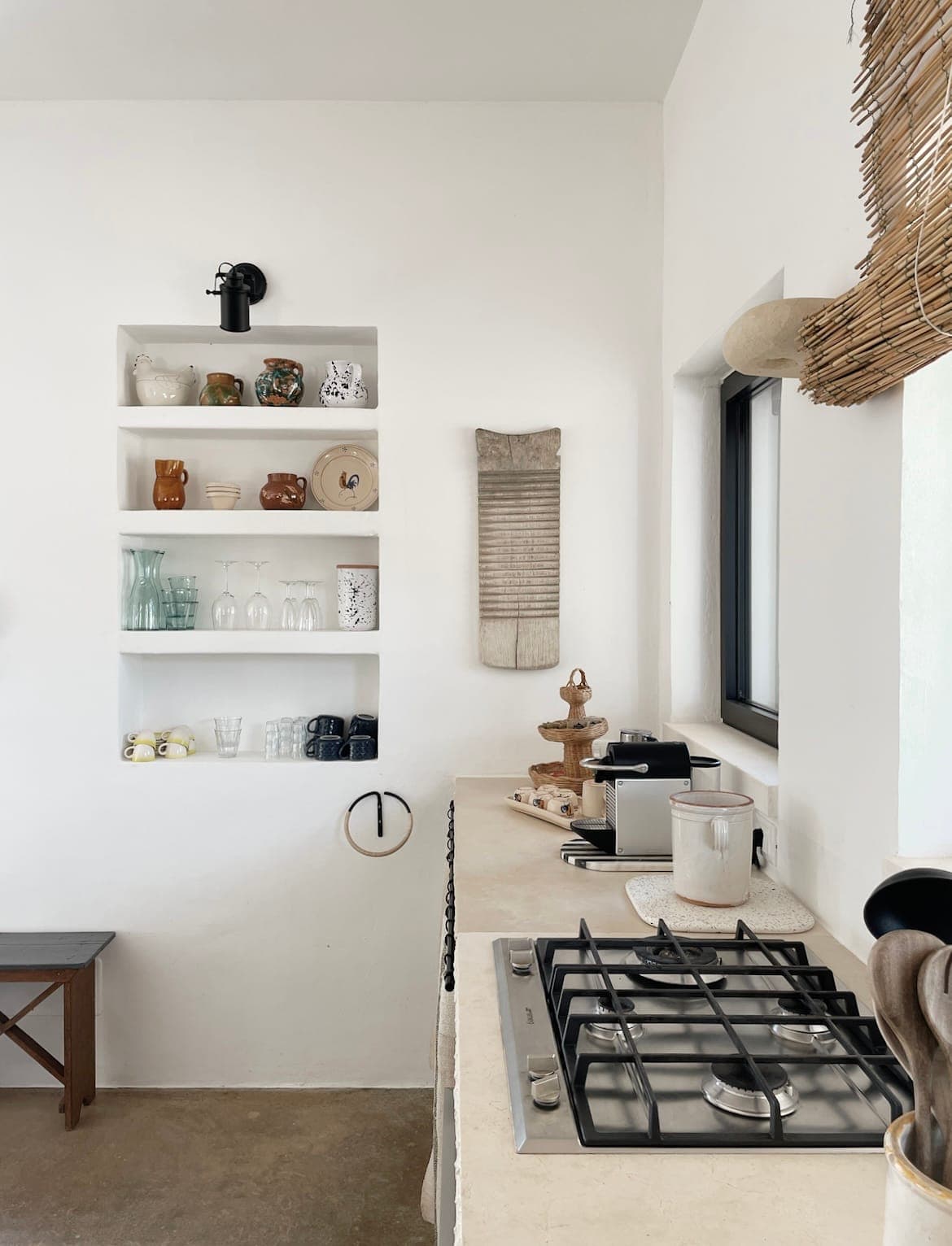 A minimalist kitchen with white walls features open shelves on which mugs and glasses are displayed. A rustic gas stove sits on a beige worktop, and a wooden bench is against the wall. A small espresso machine and decorative objects are nearby. Natural light floods in.