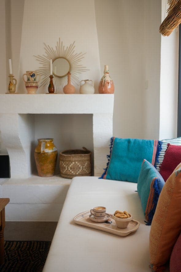 A comfortable living room with a white sofa adorned with colorful cushions. A tray with a coffee cup and cookies sits on the sofa. The background features a white fireplace with decorative pottery and a sun-shaped mirror.