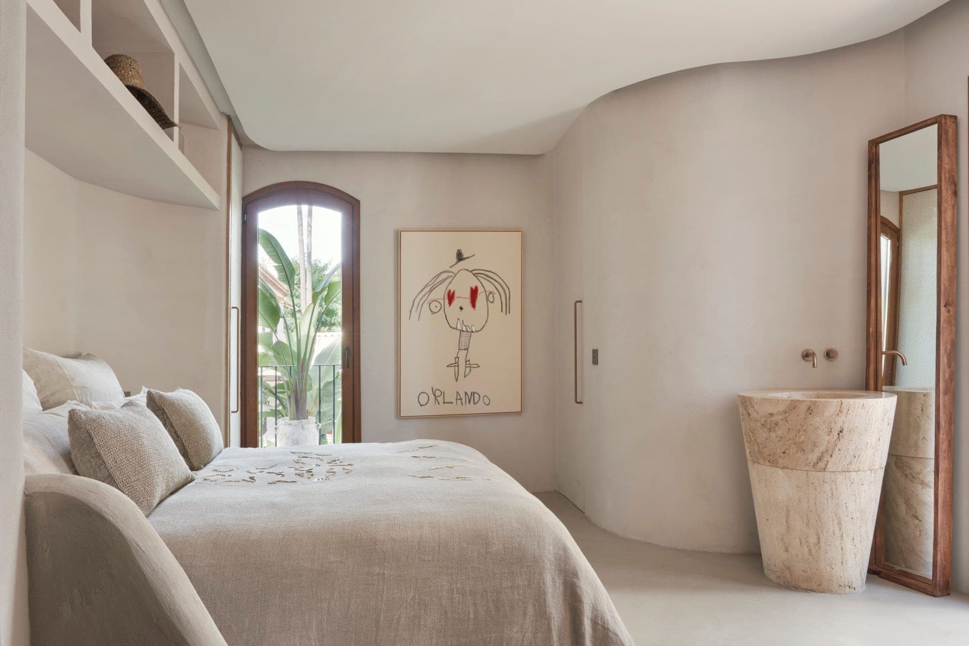 A minimalist bedroom in neutral colors features a curved wall, abstract artwork, a double bed with beige sheets, a rustic stone sink and a large mirror. A semi-curved window offers a glimpse of green foliage.
