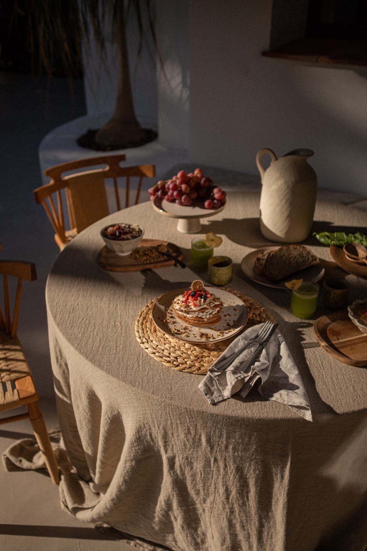 A round table, covered with a beige tablecloth, is set with a cake garnished with berries, a bowl of grapes, a pitcher, bread and two green drinks. Sunlight casts shadows, creating a warm, cosy atmosphere. Two wooden chairs are partially visible.
