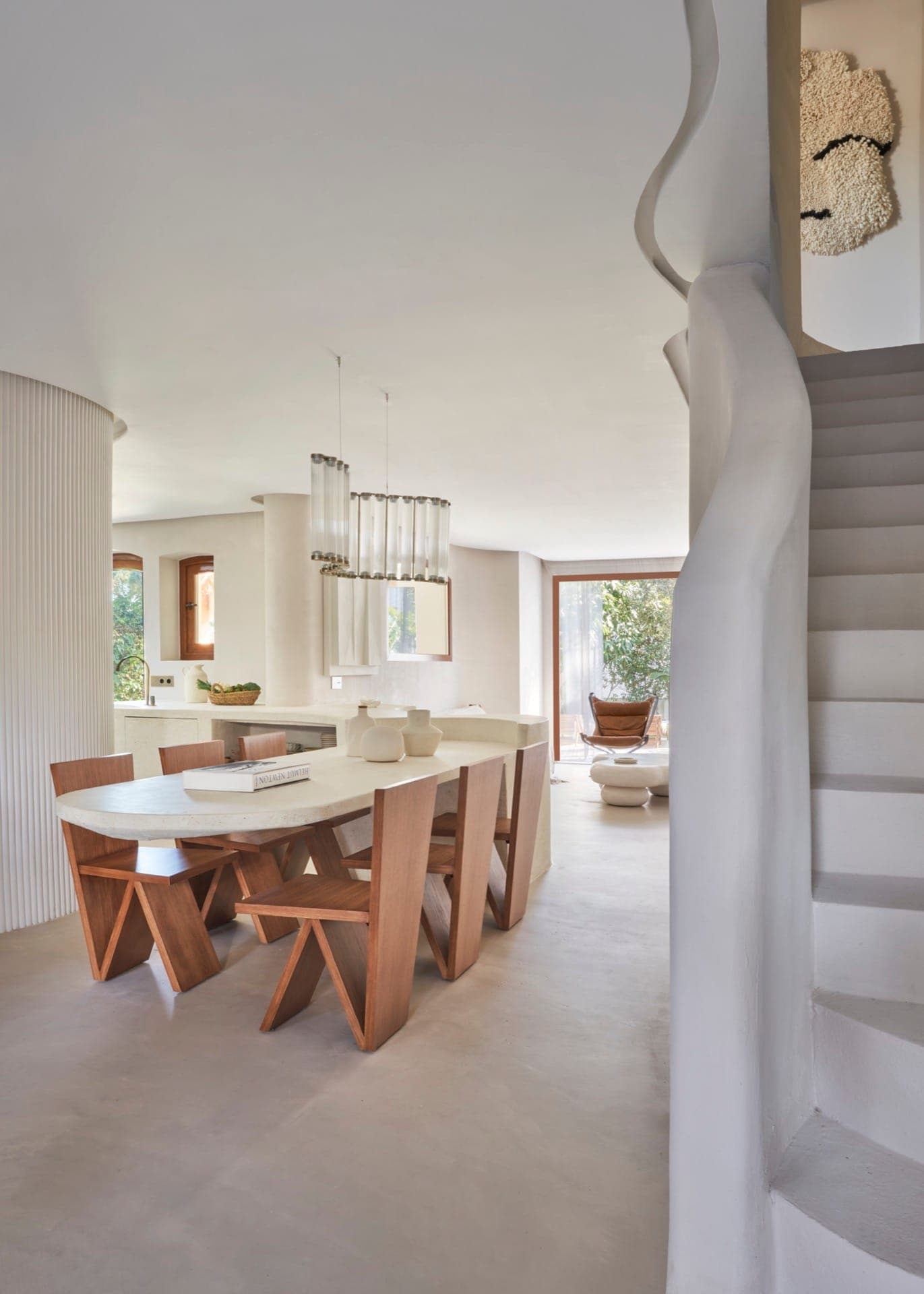 A modern dining room with a large white table surrounded by wooden chairs. Above, two contemporary hanging lamps are suspended. In the background, a window with a view of the greenery. A sculptural staircase is visible on the right.