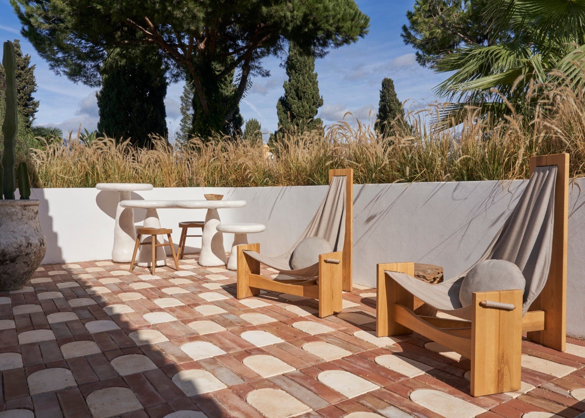 Outdoor terrace with two wooden deckchairs and a white table with stools. The floor is brick and tiled. Tall grasses and trees stand out in the background against a clear blue sky.