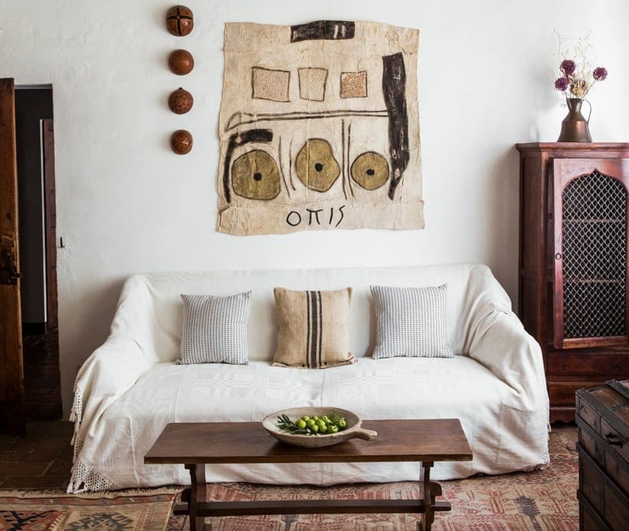 Un salon chaleureux est doté d'un canapé blanc avec des coussins à motifs et d'une table basse en bois rustique. Une grande tenture murale abstraite orne le mur blanc au-dessus du canapé. Une armoire en bois et des objets décoratifs complètent ce décor chaleureux et vintage.