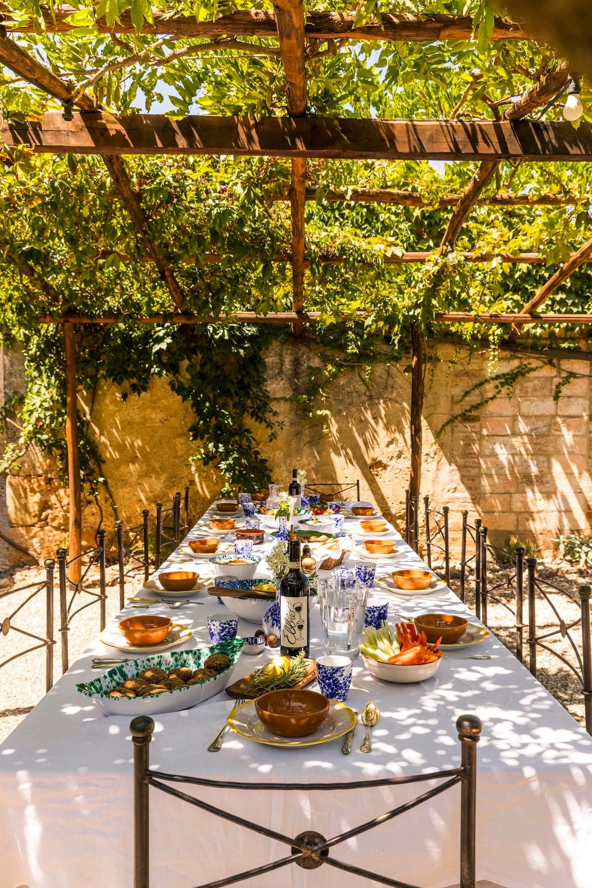 Une longue table à manger d'extérieur sous une pergola, avec de la verdure au-dessus. Elle est dressée avec une nappe blanche, de la verrerie à motifs bleus, des assiettes, des bouteilles et des couverts, dans un cadre de jardin ensoleillé et rustique.