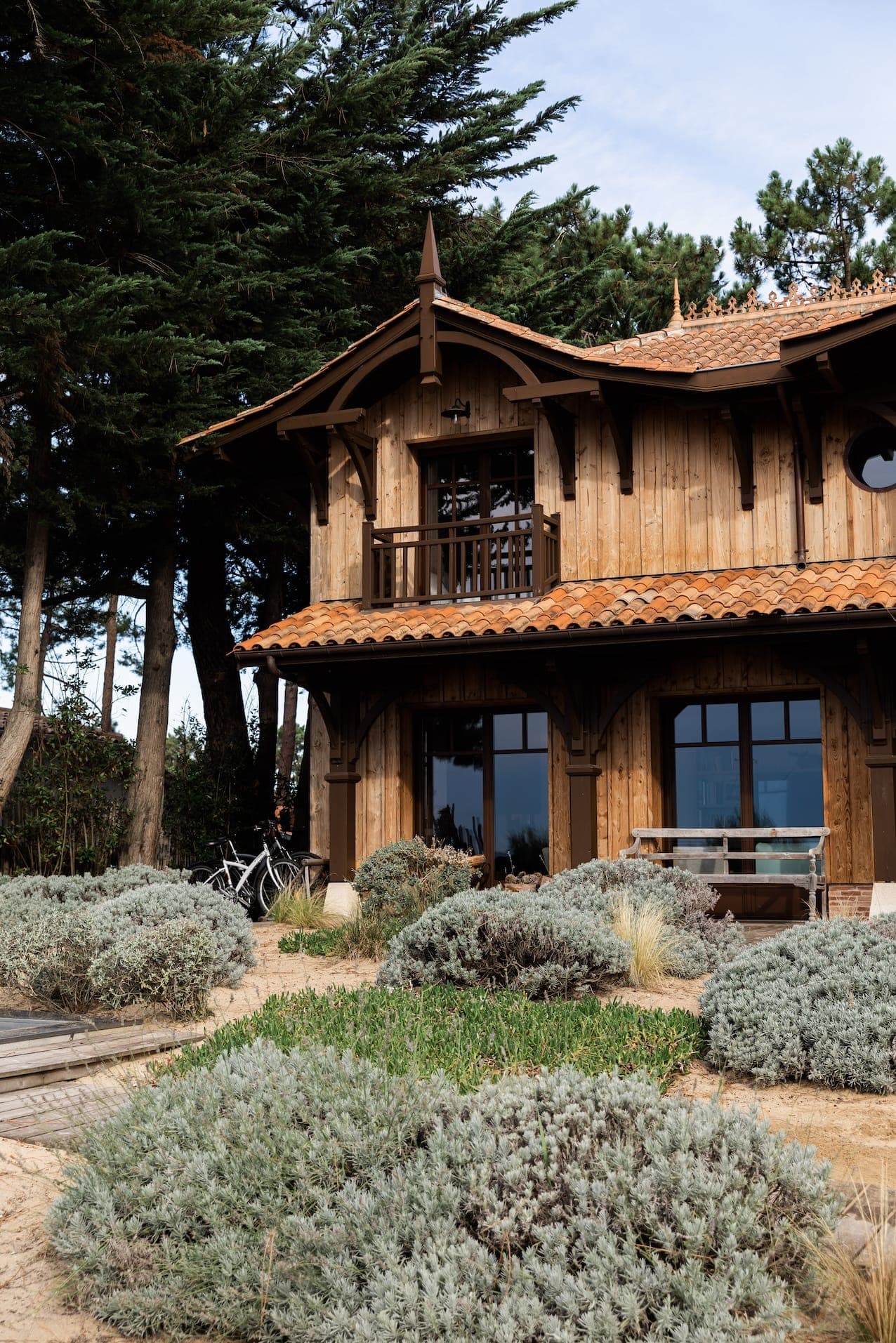 A rustic wooden house with a terracotta roof, nestled among tall trees. The first floor features large windows and lush vegetation. A bicycle is parked near the entrance, adding to the picturesque, peaceful atmosphere.