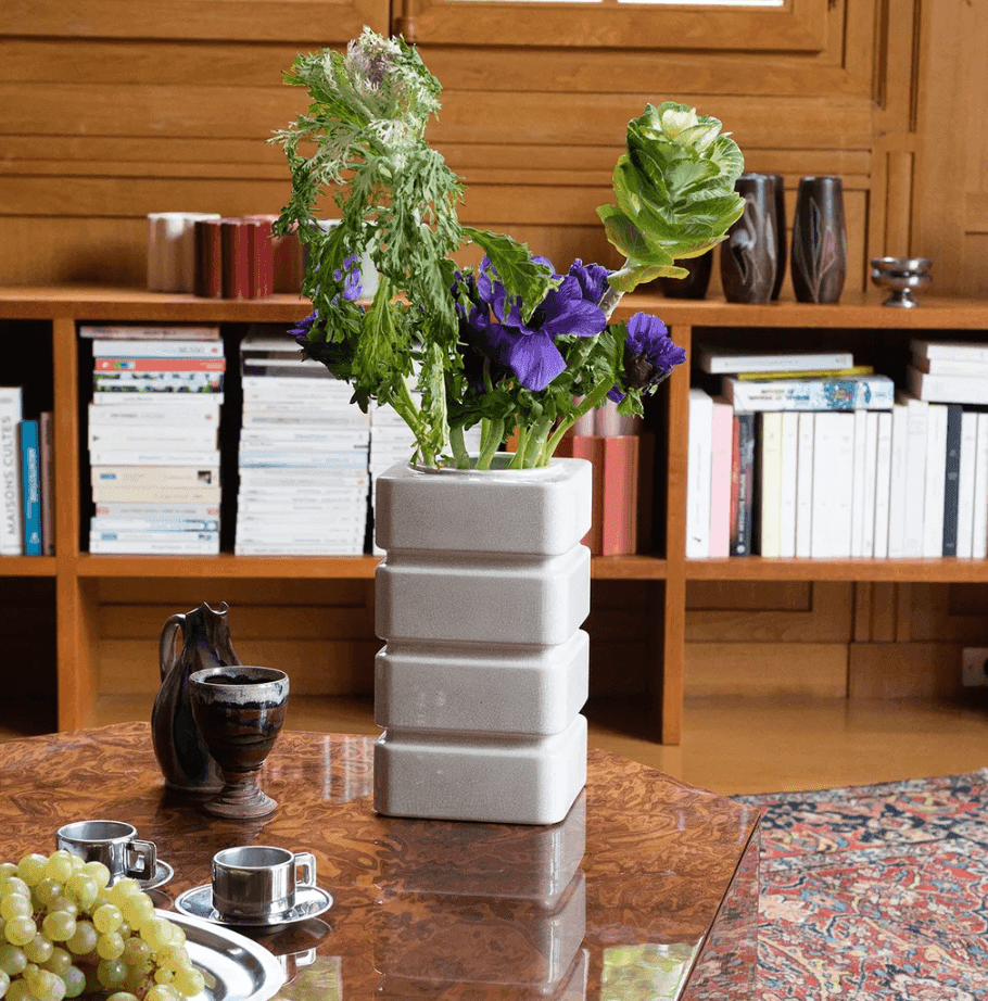 Un vase blanc moderne orné de feuillage vert et de fleurs violettes est posé sur une table en bois. À proximité se trouvent deux tasses à expresso, un petit pichet et une assiette de raisins. Une bibliothèque bien garnie se trouve à l'arrière-plan.