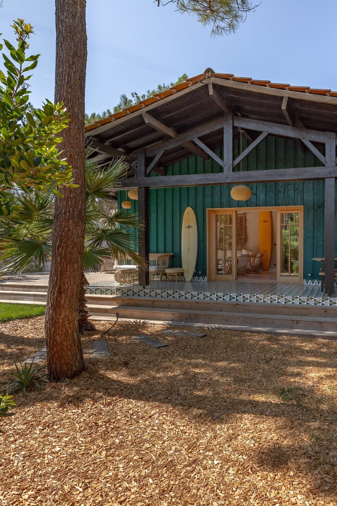 A rustic wooden cabin with a teal-blue facade and red-tiled roof, surrounded by trees. The porch features surfboards, wicker furniture and large windows. The floor is covered with wood shavings and the sky is clear.