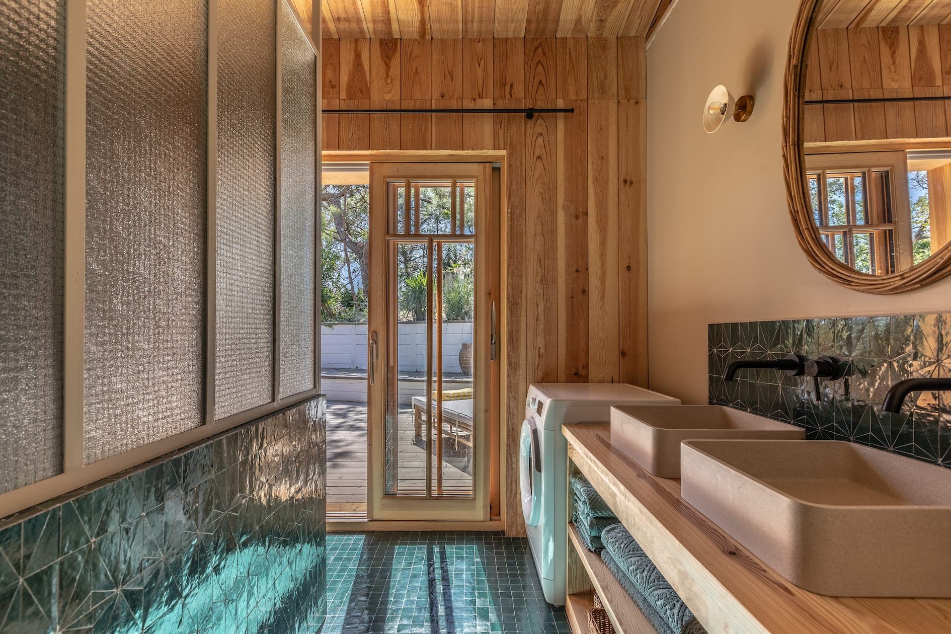 A modern bathroom with wood-panelled walls and ceiling, featuring a double vanity with round mirrors, washing machine and green textured tiles. Sunlight filters through a glass door to the outside.
