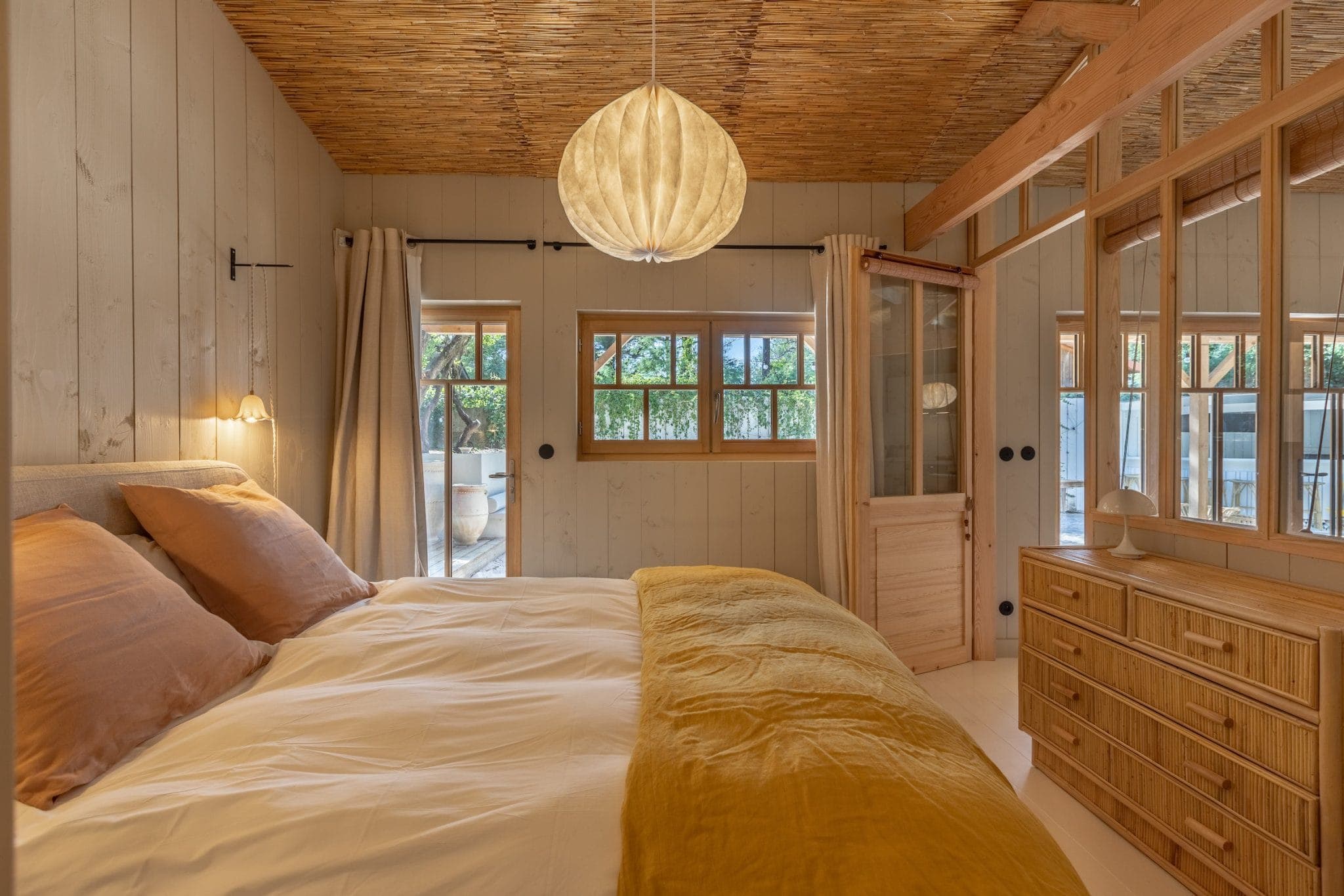 Comfortable wooden bedroom with king-size bed, decorated with brown and white linen. A spherical lamp hangs from the ceiling. Windows let in natural light; a sliding door leads outside. A wooden chest of drawers is on the right. Earthy tones create a warm ambience.