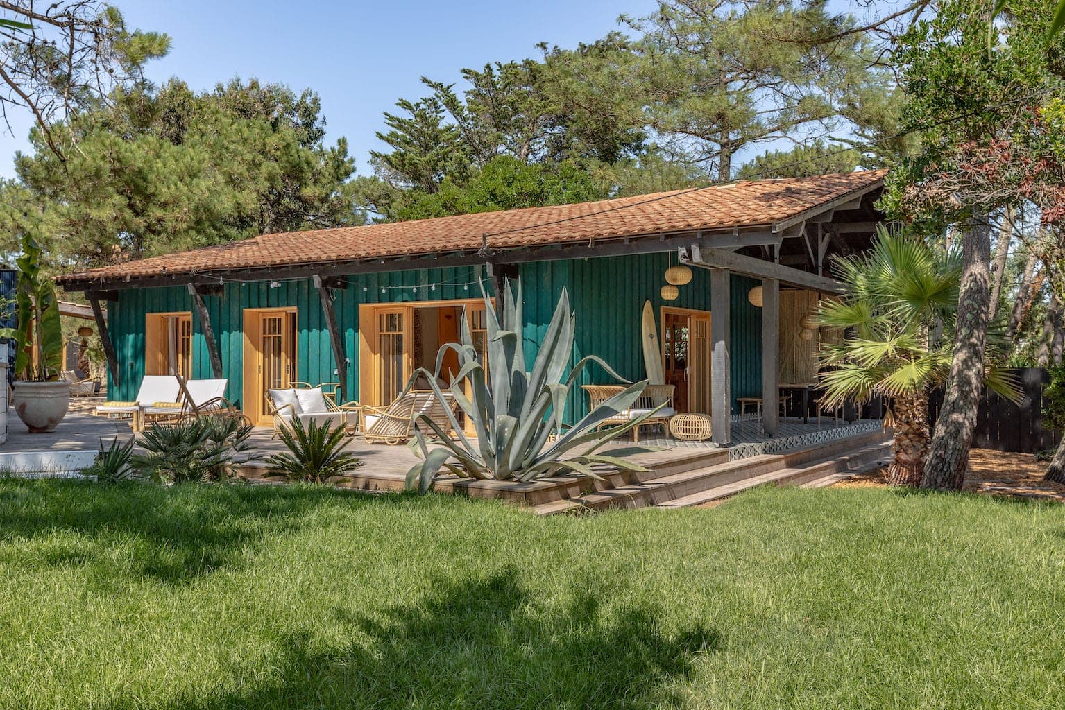 A green wooden house with a red-tiled roof is surrounded by lush grass and various plants. It features multiple wooden patio doors, outdoor seating, and is set amidst trees on a sunny day.