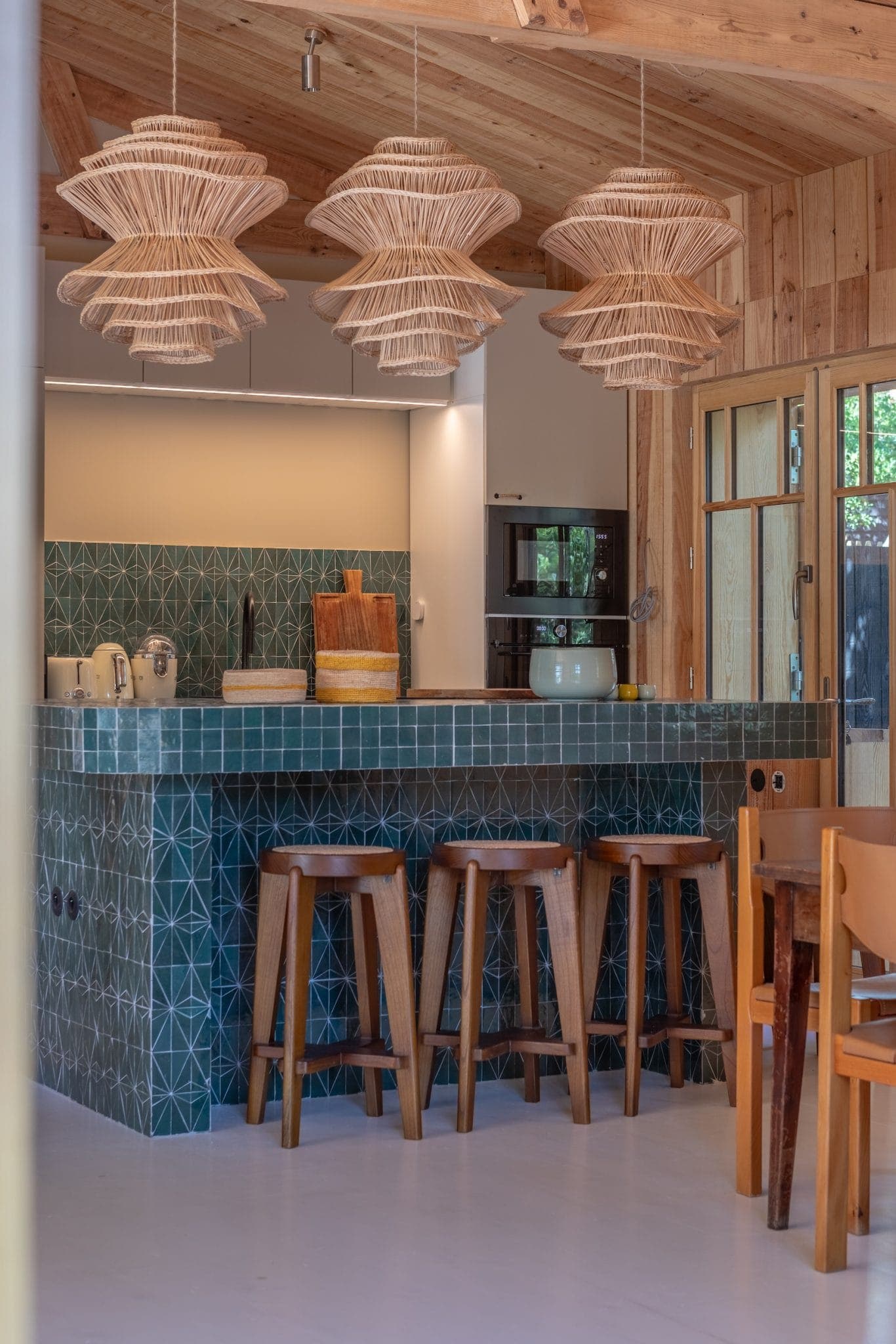 A rustic kitchen with a green-tiled island, wooden stools and wicker pendant lights. The background features a wooden ceiling and walls, modern appliances and a large window to let in natural light.