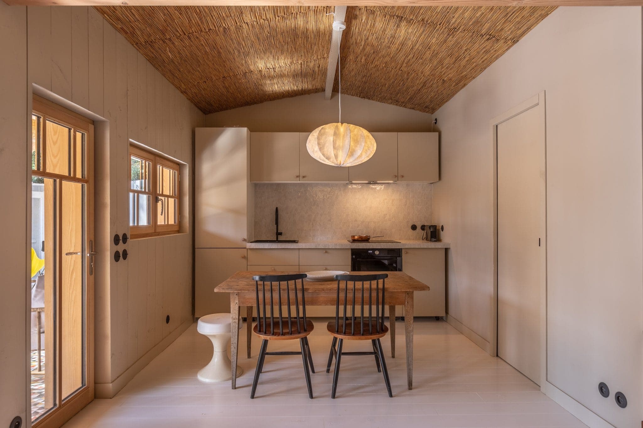 A comfortable kitchen with a wooden table and two chairs. Light wood cabinets and countertops, modern appliances and a warm ceiling light add to the welcoming atmosphere. A window and door let in natural light, and a rustic woven ceiling completes the look.