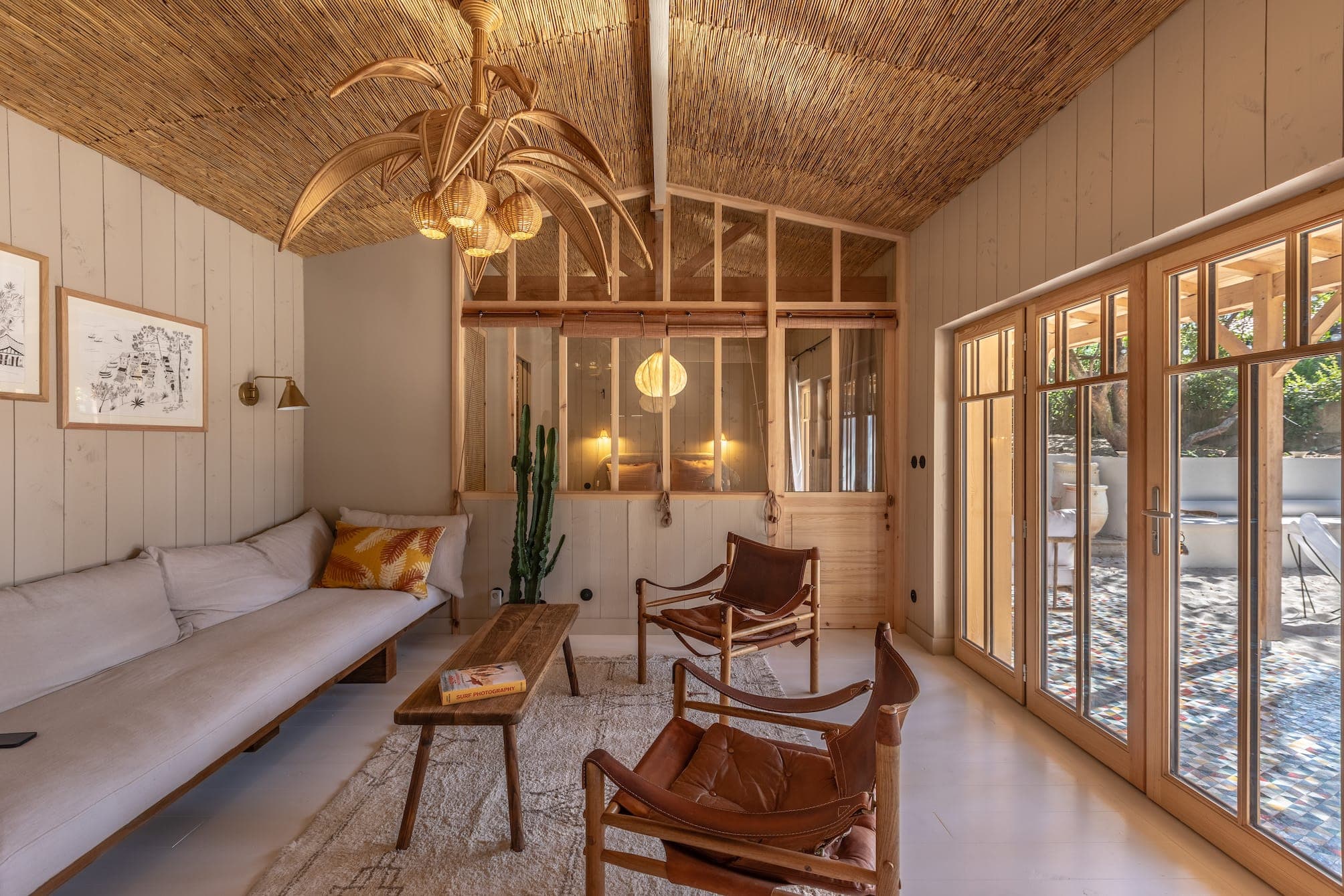 A comfortable living room with a white sofa, two brown leather chairs and a wooden coffee table. The ceiling is woven fabric and a unique chandelier hangs in the center. Large windows allow natural light to fill the space, highlighting a potted cactus.
