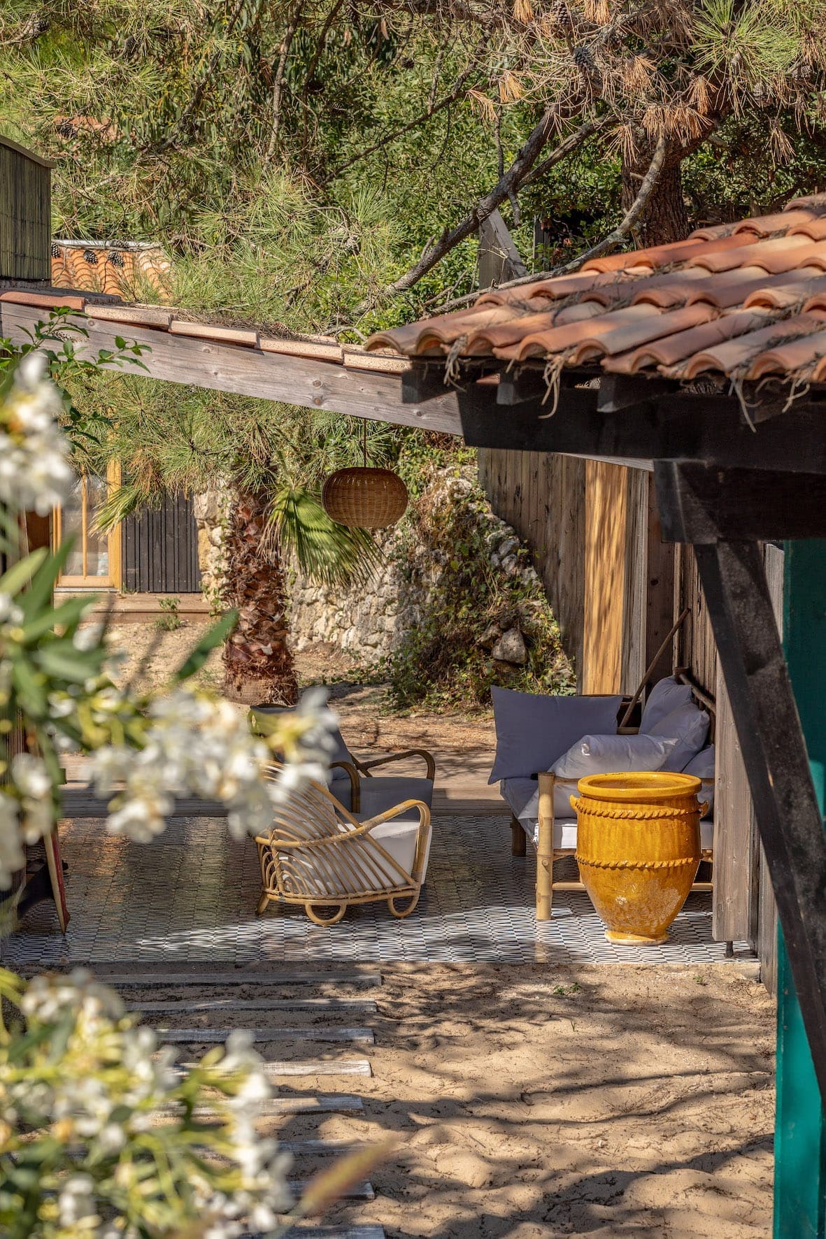 Espace extérieur cosy avec chaises en osier, table d'appoint jaune et coussins, sous une pergola en bois. Entouré de verdure et de sable, le toit en tuiles d'argile est partiellement visible.