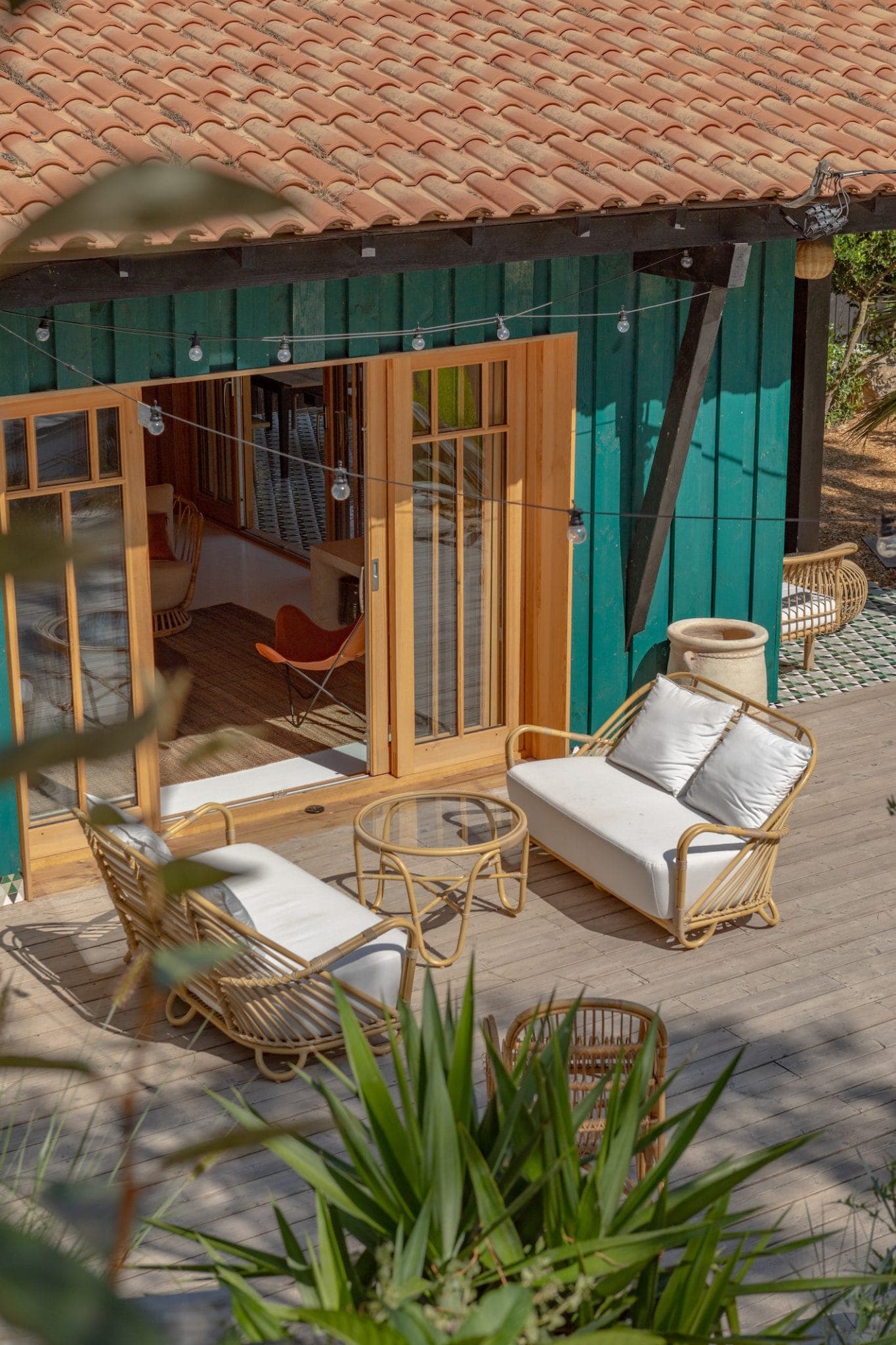 A comfortable outdoor seating area with two rattan sofas and a round glass coffee table on a wooden terrace. The terrace is attached to a green wooden house with large glass doors. Strings of lights hang overhead and greenery surrounds the space.