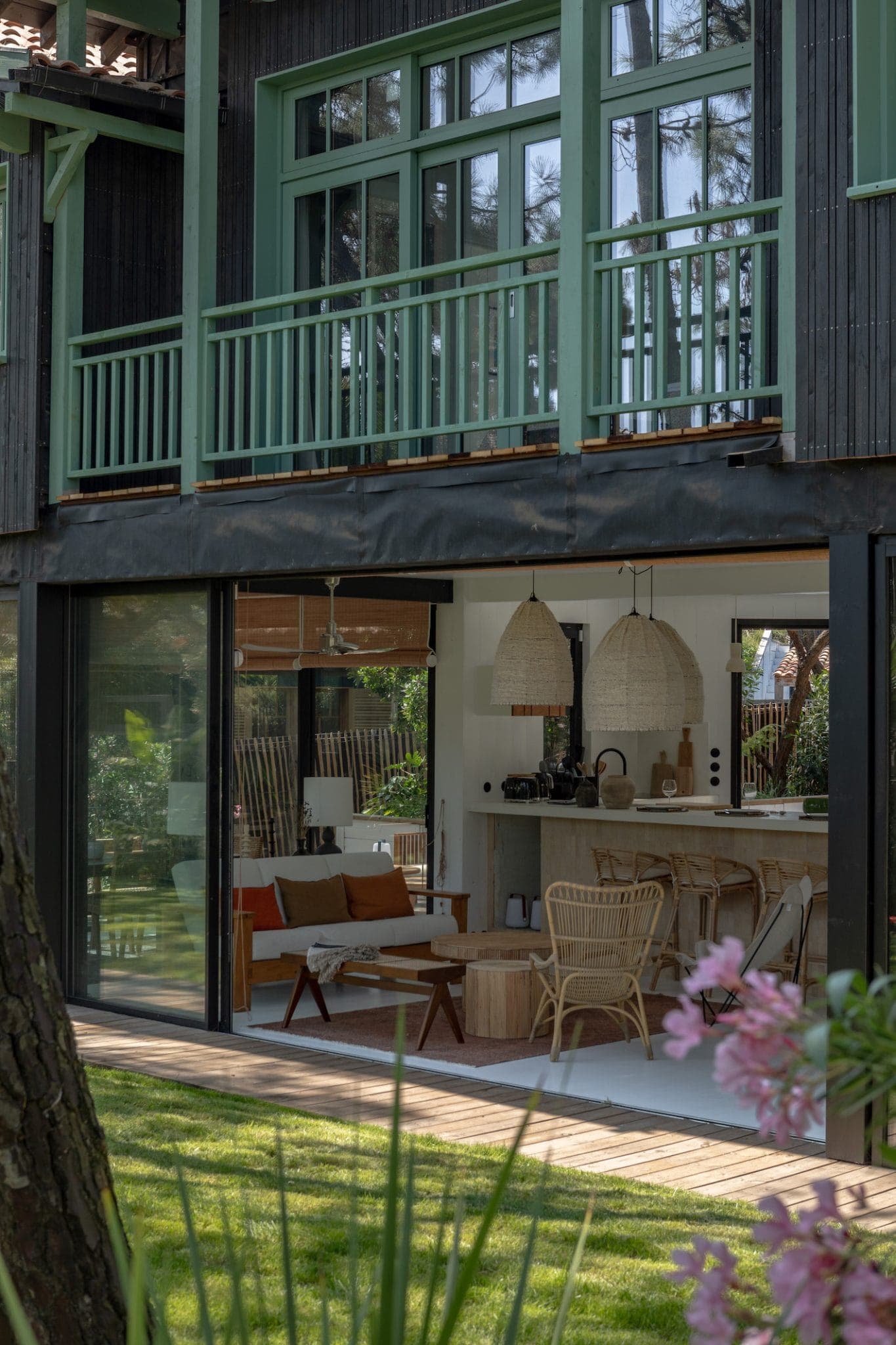 A modern home with large glass doors that open to reveal a welcoming living space. Inside are wicker chairs, a rustic table and wicker hanging lights. The exterior features a wooden terrace and lush garden with trees and blossoming flowers in the foreground.