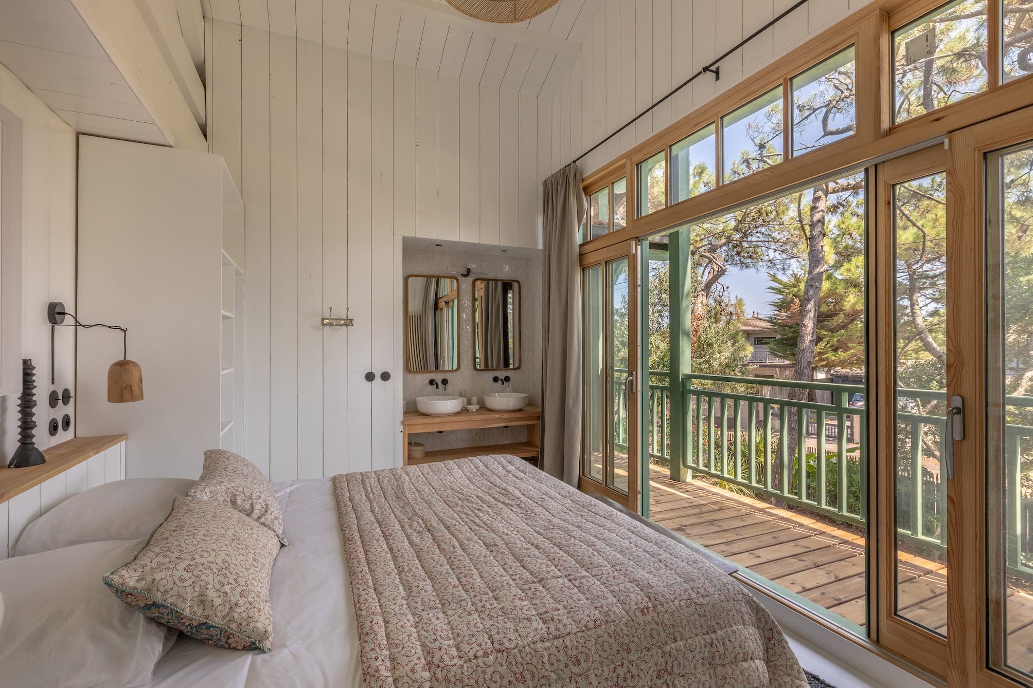 A comfortable bedroom with white wooden walls features a double bed with patterned bedding, a wooden dressing table with twin sinks and mirrors, and large glass doors opening onto a balcony overlooking the trees. Natural light fills the room.