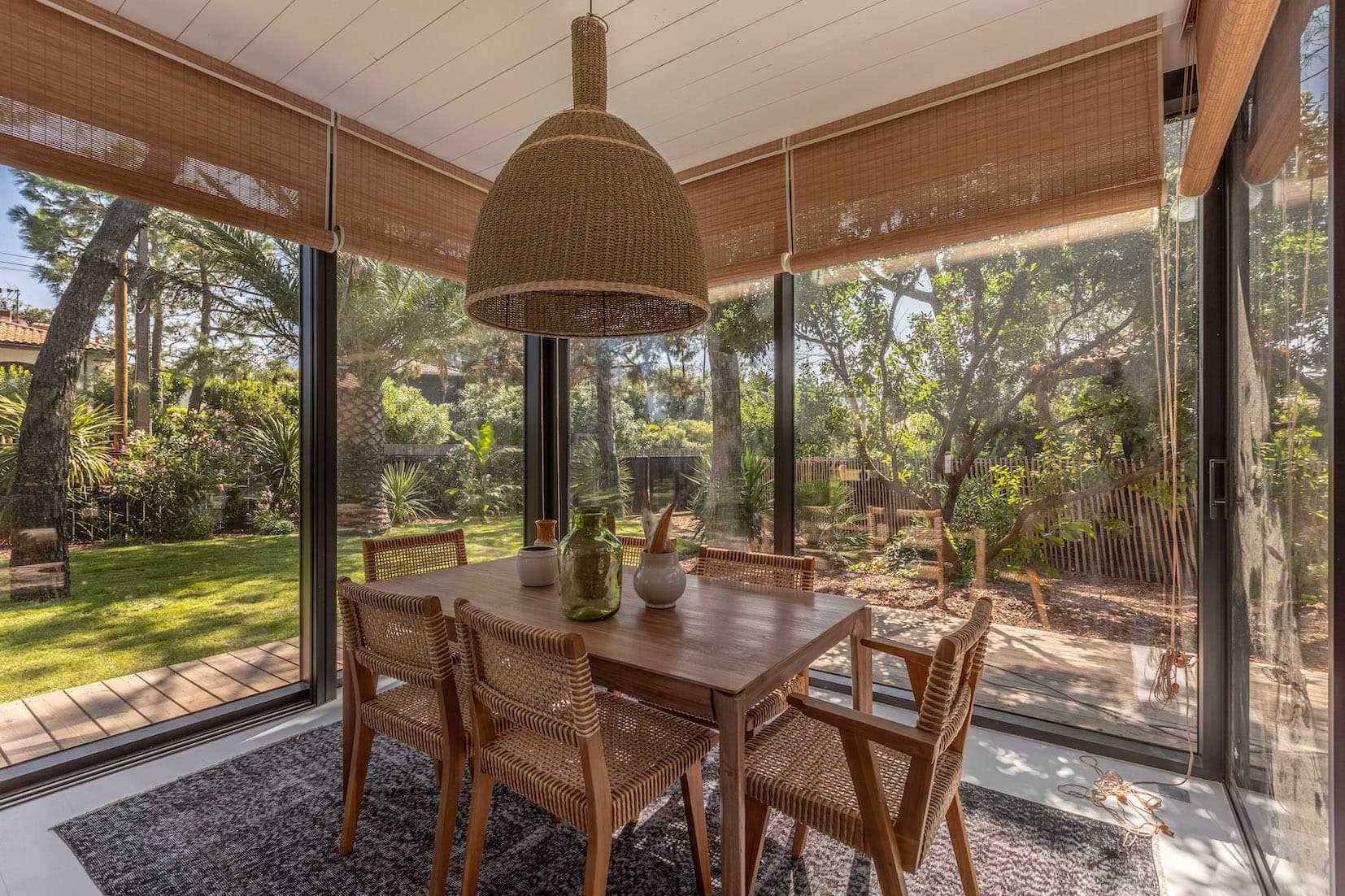 A bright veranda with large windows offering views of the lush garden. The room features a wooden dining table with six wicker chairs and a woven pendant light. Natural light filters in through bamboo blinds, creating a warm and welcoming atmosphere.