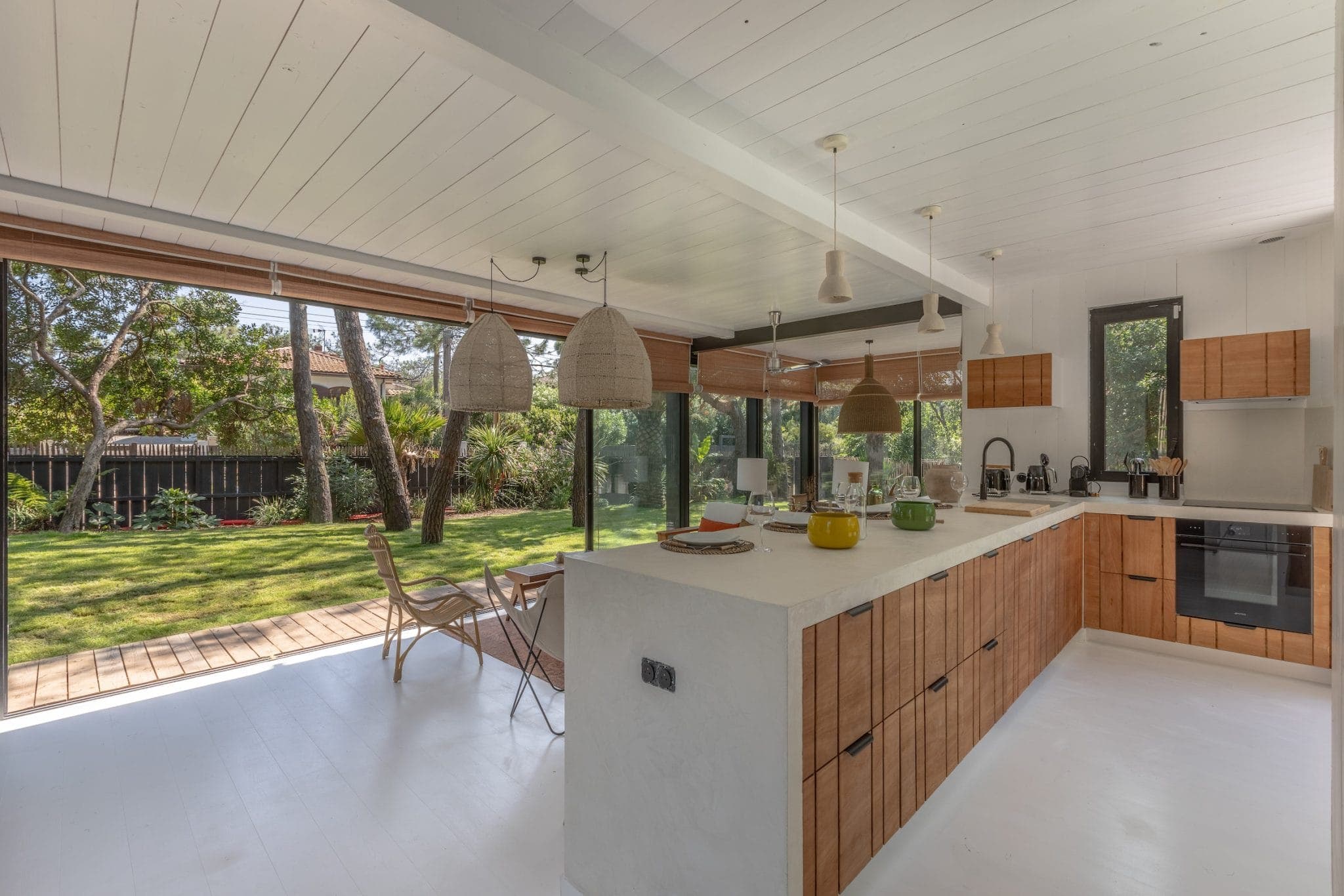 A modern kitchen with wood cabinets and a white island. Large windows offer a view of a leafy backyard. Pendant lights hang over the island and two rattan chairs face the windows. The space is bright and airy.