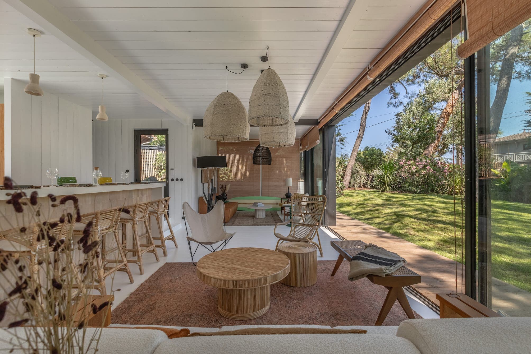 A modern living room with large windows offering garden views. It features wicker hanging baskets, a wooden coffee table, bar stools and a patterned rug. The outdoor landscape with trees and greenery is visible through the glass doors.