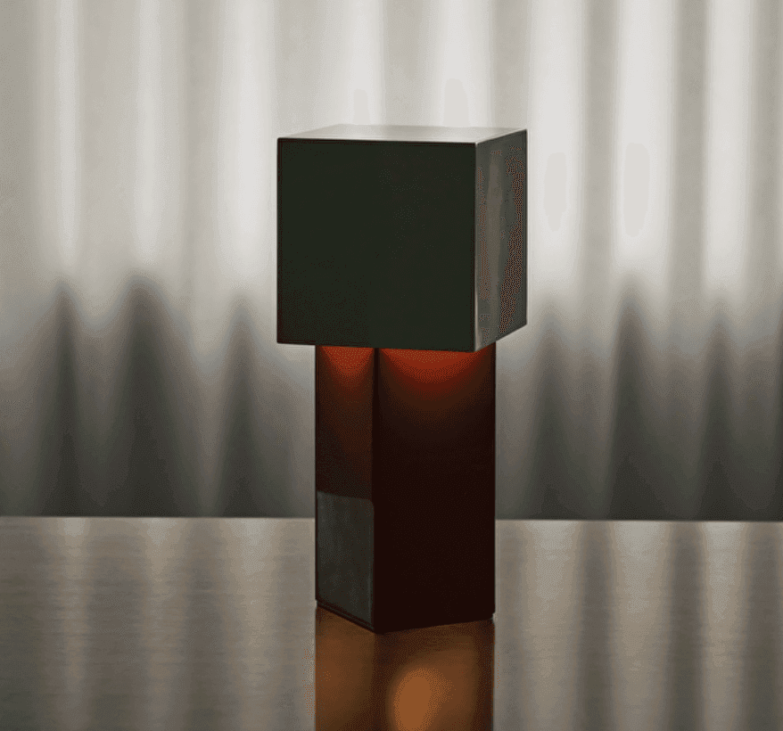 Une lampe carrée moderne et minimaliste, dotée d'un abat-jour vert foncé et d'une base sombre, repose sur une surface réfléchissante. Une douce lumière jaillit de l'intérieur, projetant des ombres subtiles sur un fond gris flou.