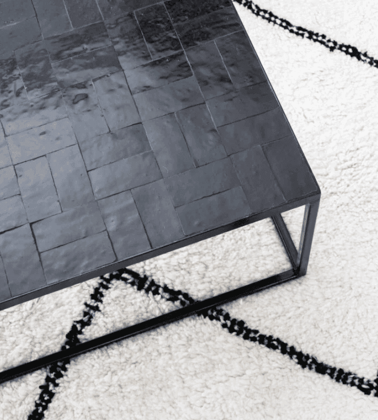 Une table basse noire brillante, ornée d'un motif de carrelage, est posée sur un tapis blanc aux motifs géométriques noirs. La table est dotée d'une structure métallique simple et sa surface reflète subtilement la lumière.
