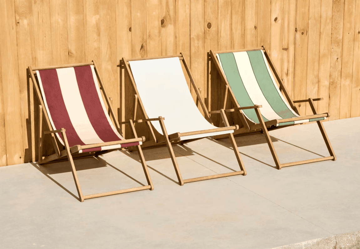 Trois chaises longues en bois sont disposées sur une terrasse ensoleillée, contre une clôture en bois. Les chaises sont dotées de tissus rayés marron et beige, beige uni et vert et beige.