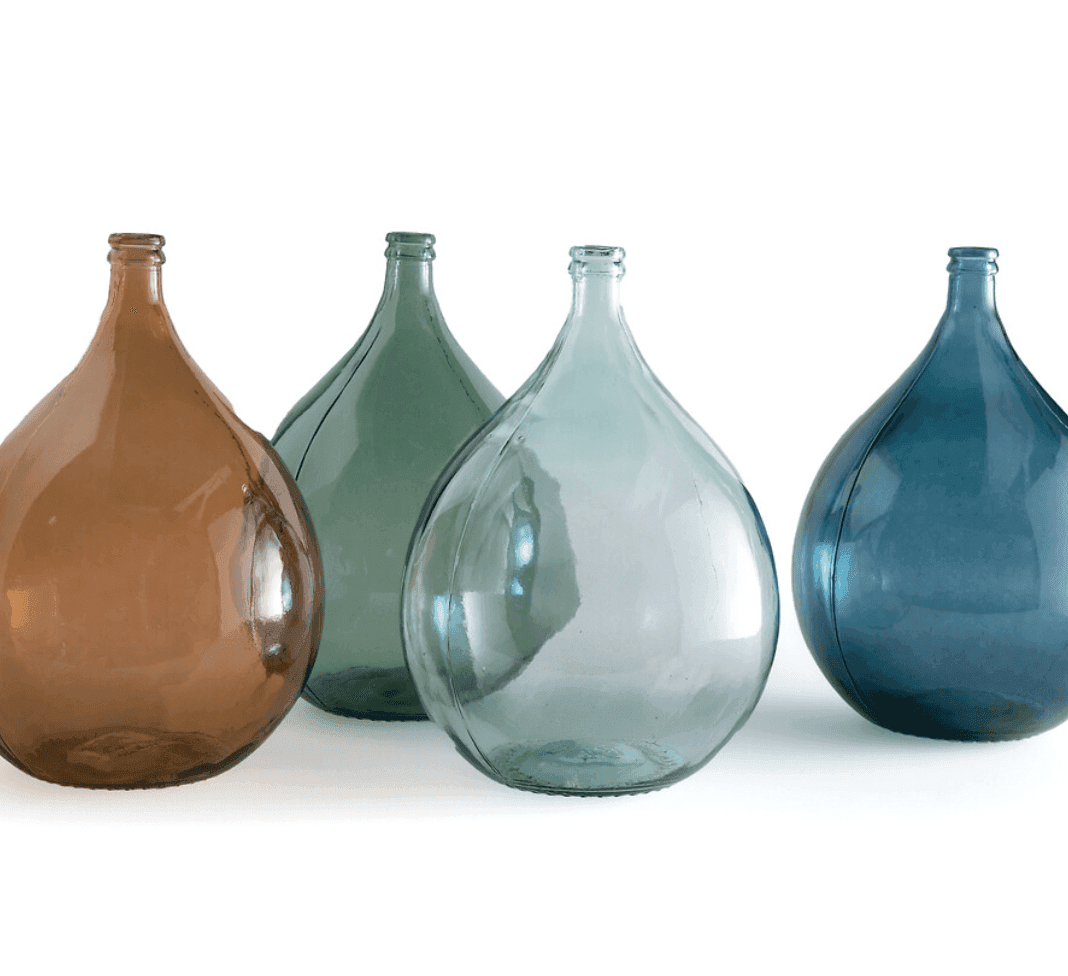 Quatre grandes bouteilles en verre en forme de larme aux teintes translucides variées (ambre, vert, transparent et bleu) sont disposées en rangée sur un fond blanc.
