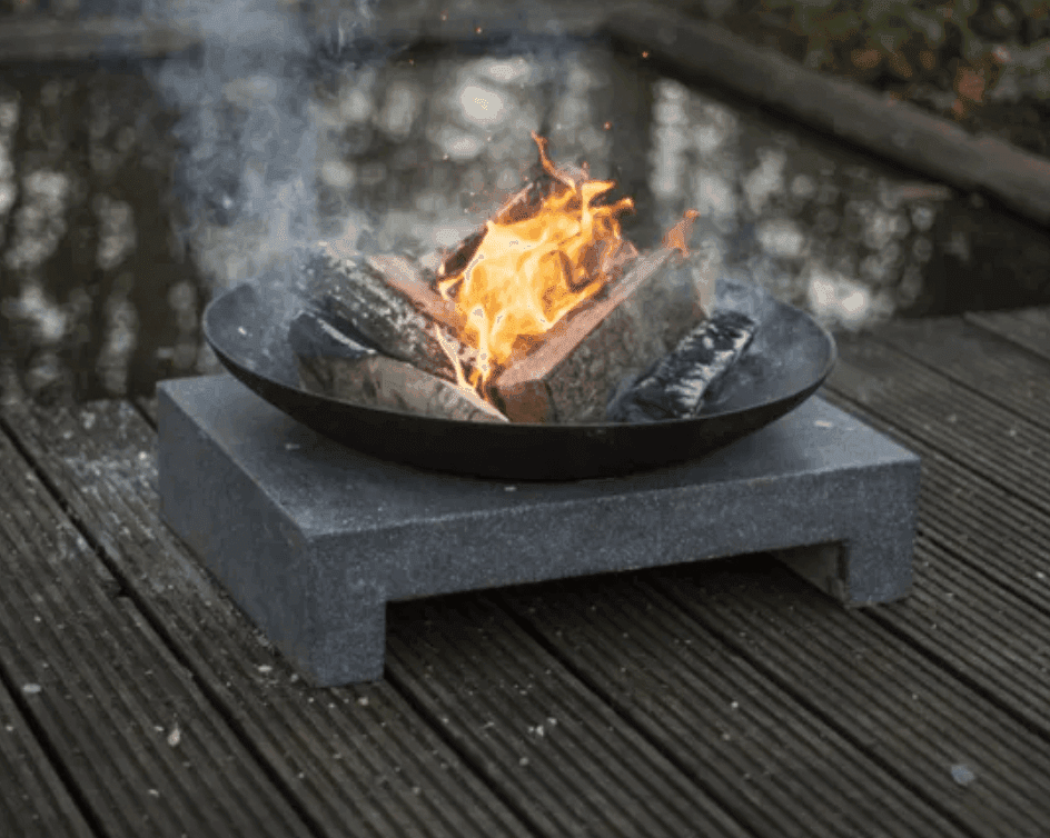 Un foyer avec des bûches enflammées est installé sur une plate-forme en pierre. De la fumée s'élève des flammes. Le foyer est placé sur une terrasse en bois et un élément aquatique réfléchissant est partiellement visible en arrière-plan.