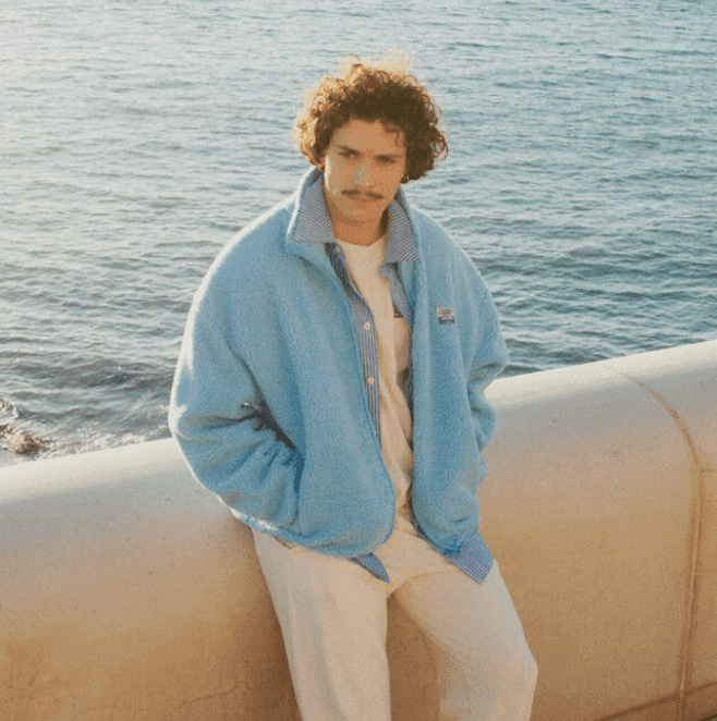 Un homme aux cheveux bouclés et à la moustache, vêtu d'une veste bleu clair et d'un pantalon blanc, s'appuie contre un mur de pierre au bord de la mer. L'océan est calme et le soleil se couche, projetant une lumière chaude sur la scène.