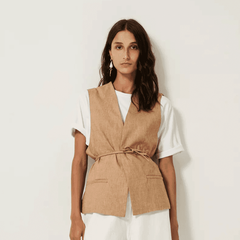 Femme portant un gilet beige sur un t-shirt blanc et un pantalon blanc. Elle se tient debout sur un fond uni et clair. Ses cheveux sont ondulés et elle porte une boucle d'oreille pendante.
