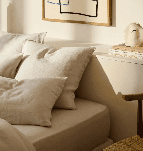 Un coin chambre douillet avec un lit aux oreillers et draps beiges et blancs moelleux. Une petite table de nuit en bois contient des livres empilés et un objet de décoration fantaisiste en forme de chat. Au-dessus, une œuvre d'art abstraite encadrée est accrochée au mur.