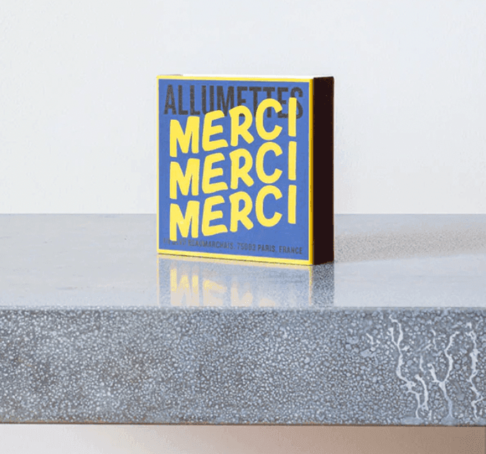 Une boîte carrée d'allumettes portant l'inscription « ALLUMETTES MERCI MERCI MERCI » en lettres jaunes audacieuses sur fond bleu repose sur une surface grise mouchetée. Au fond de la boîte, on peut lire « 111 Boulevard Beaumarchais, 75003 Paris, France ».
