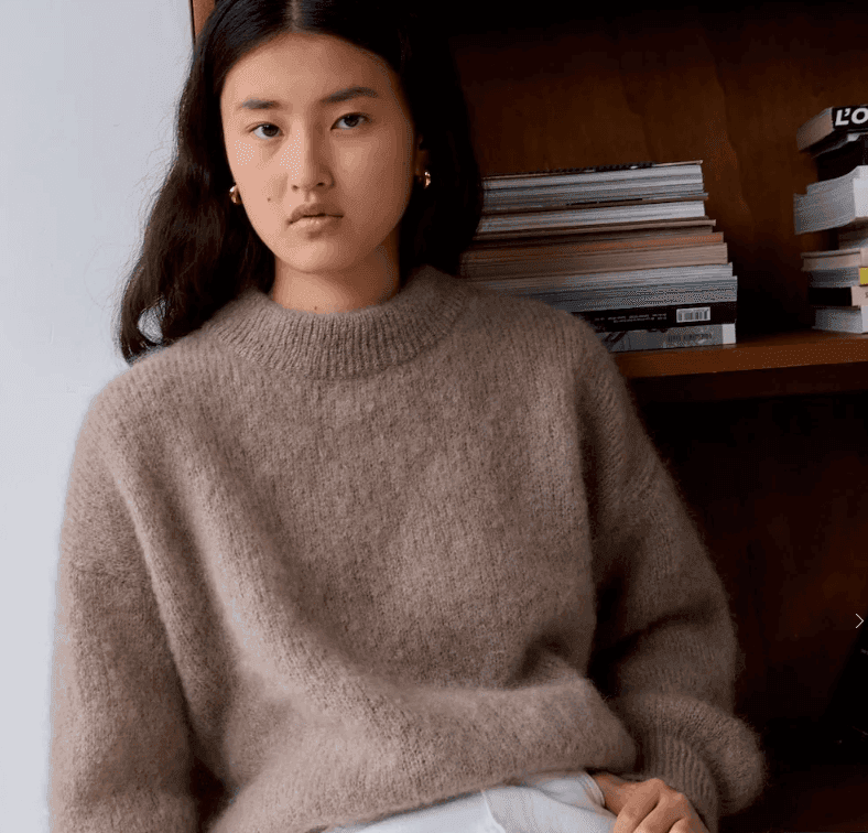 Une personne aux cheveux longs et foncés porte un pull marron confortable et surdimensionné et un pantalon blanc, assise devant une étagère en bois remplie de livres empilés. Elle a une expression neutre et regarde directement la caméra.