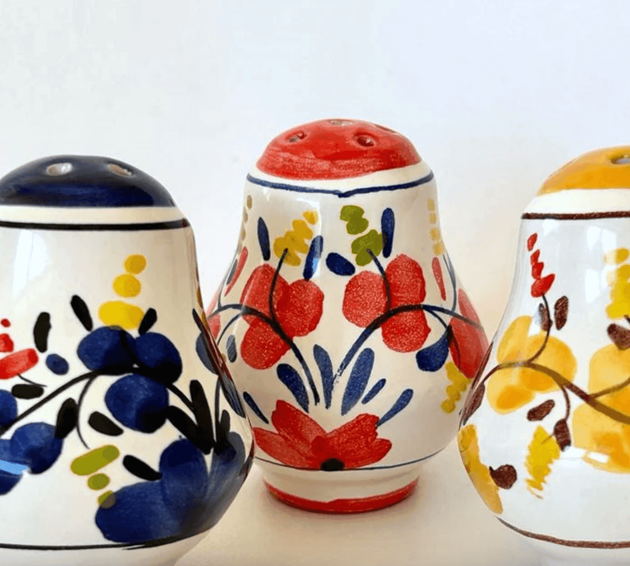 Trois shakers en céramique aux motifs floraux. Celui de gauche est bleu avec des motifs jaunes et noirs, celui du centre est rouge avec des touches de vert et de jaune, et celui de droite est jaune avec des détails rouges et marron, le tout sur fond blanc.