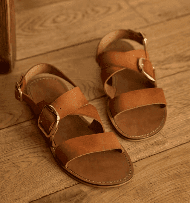 Une paire de sandales en cuir marron à larges brides et à grandes boucles métalliques est posée sur un parquet. Leur design est simple et rustique.