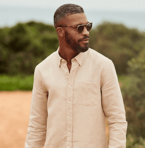 Une personne portant des lunettes de soleil et une chemise beige se tient dehors, avec un feuillage vert flou en arrière-plan. Elle a les cheveux courts et une barbe rasée, et regarde de côté.