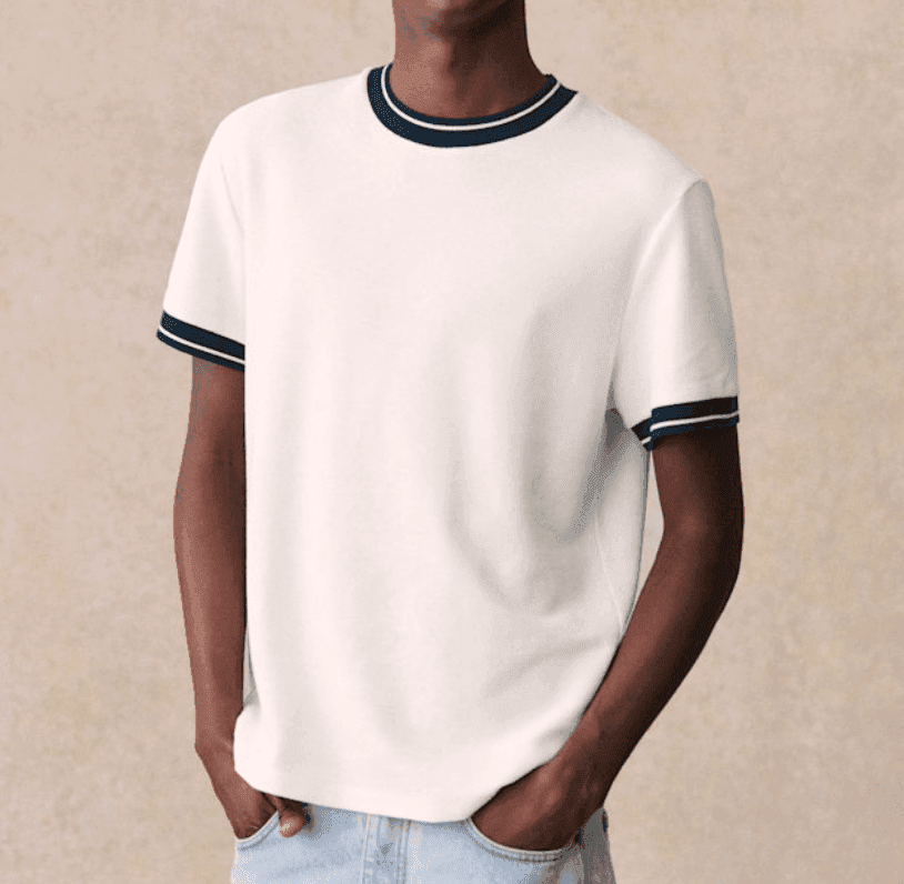 Une personne vêtue d'un t-shirt blanc avec un liseré noir au col et aux manches se tient debout, les mains dans les poches d'un jean bleu clair. L'arrière-plan est uni, beige clair. La tête n'est pas visible sur l'image.