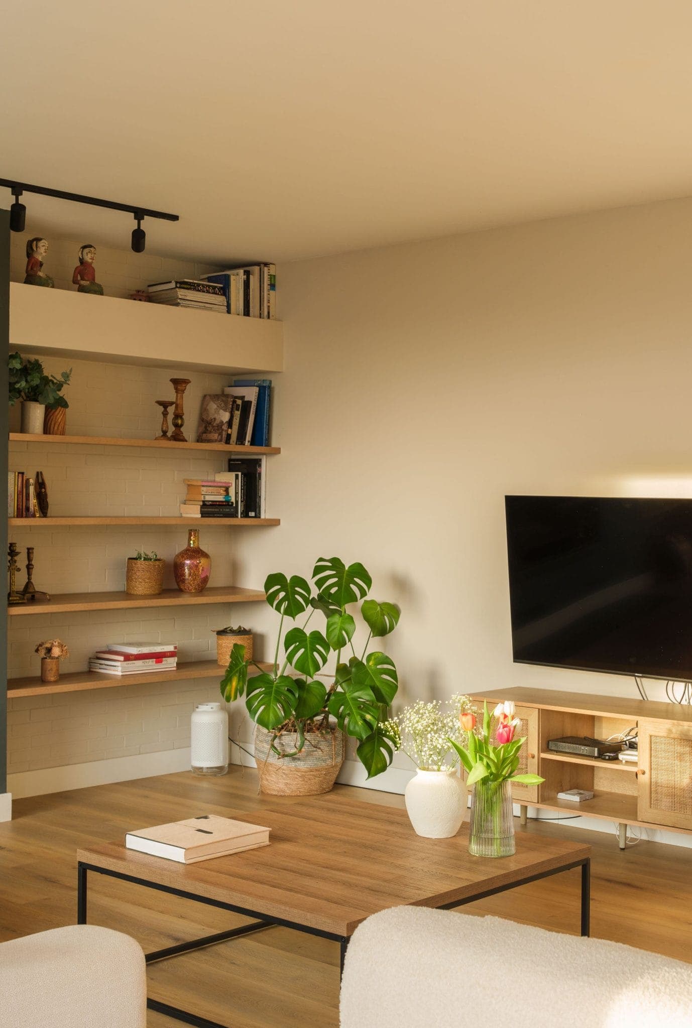 A cozy living room with a wall-mounted TV, wooden shelves for books and decorative objects, and a coffee table adorned with plants and flowers. The space, with its light-colored walls and comfortable sofa, creates a warm atmosphere.