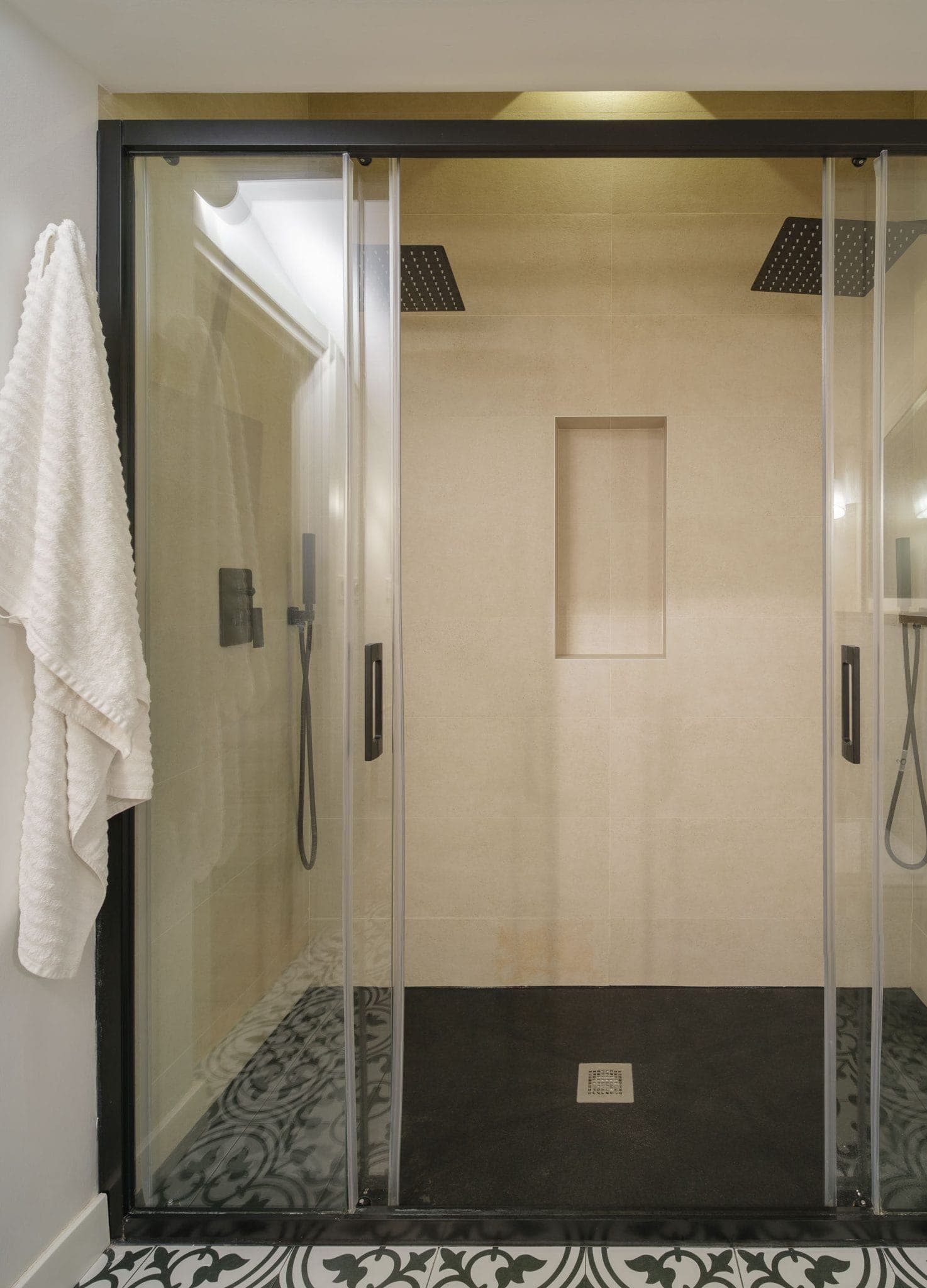 Modern shower with clear glass sliding doors, beige tiles, double rainfall showerhead and integrated niche. A white towel hangs from a black hook on the outside. The floor is tiled with black and white patterns.