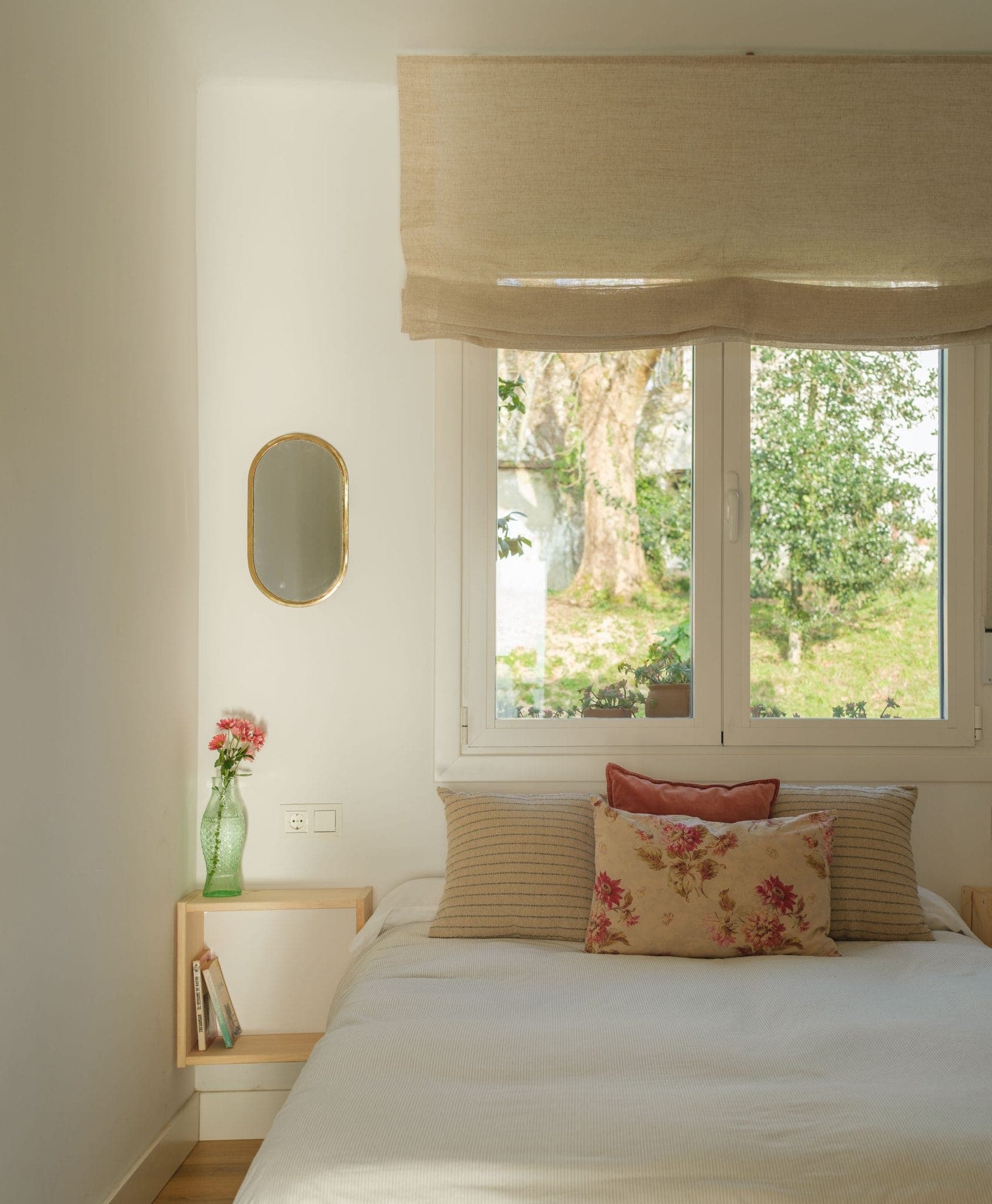 A cosy room with a carefully made bed, decorated with beige and flowery cushions. A small wooden shelf beside the bed houses a green vase adorned with pink flowers, and a mirror hangs on the wall. A window with beige curtains offers a view of the greenery outside.