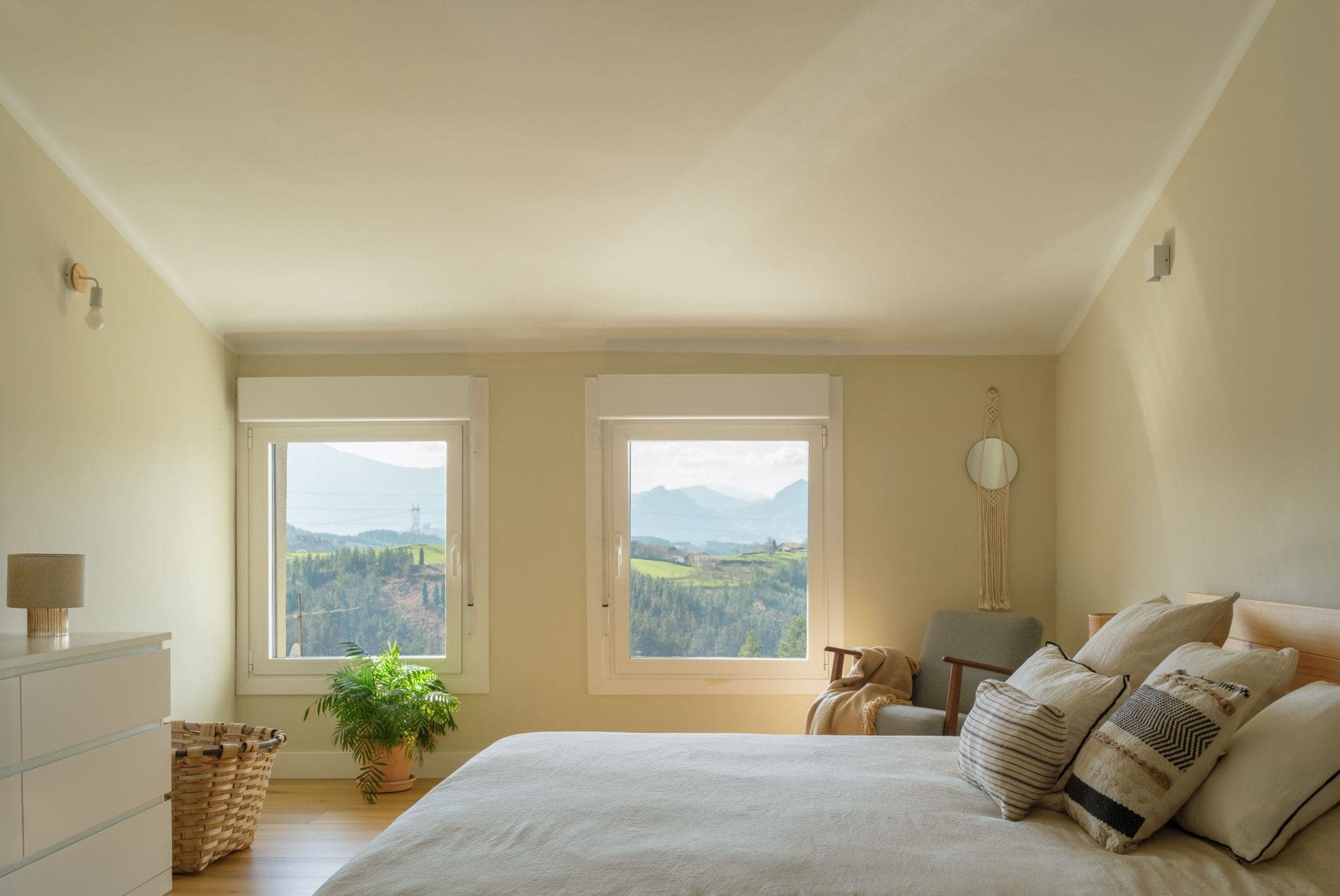 A warm room with light-colored walls features a bed with patterned pillows, a small chest of drawers and a basket. Large windows offer panoramic views of the mountains and greenery. A plant and decorative objects complete this serene setting.