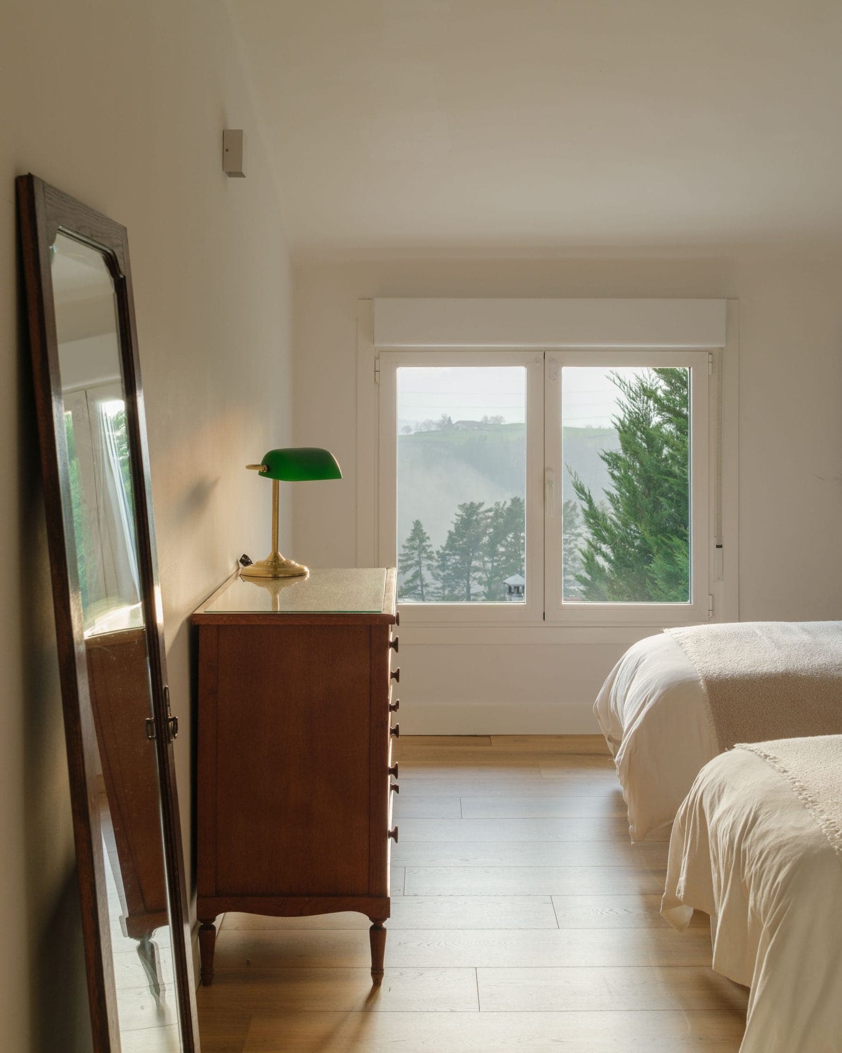 A minimalist bedroom with two beds and white bedding. A wooden chest of drawers with a green lamp stands next to a large window overlooking the trees. A large mirror leans against the wall, letting in the sunlight.