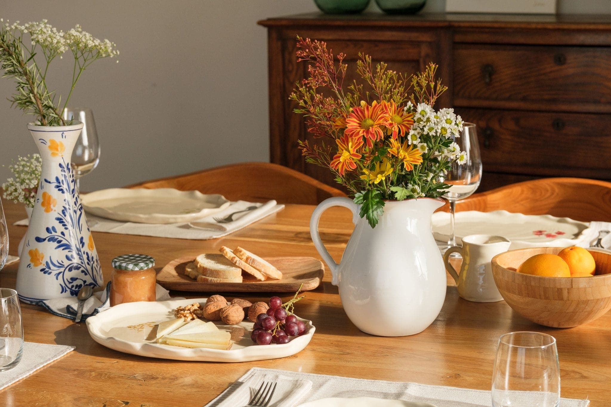 A dining table with a floral arrangement in a white pitcher, another vase with flowers, a wooden bowl of oranges, a cheese platter with grapes and nuts, a small jar of spread and wine glasses, all set on a wooden buffet.