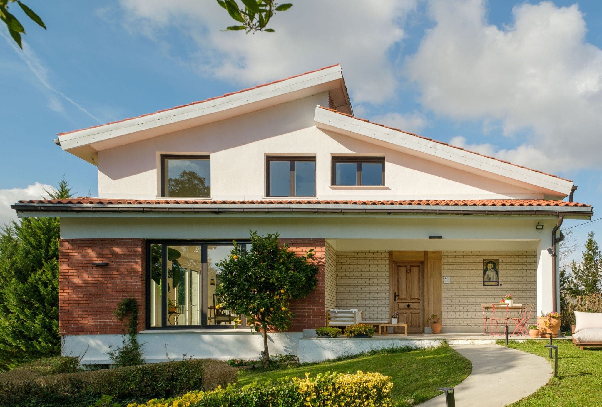 Une maison moderne à deux étages avec un toit en pente, des murs blancs et une façade en briques rouges. Elle comprend un petit porche avec une porte en bois, quelques chaises et une table. Le paysage est bien entretenu, avec des arbustes verts et un arbre. Le ciel est partiellement nuageux.